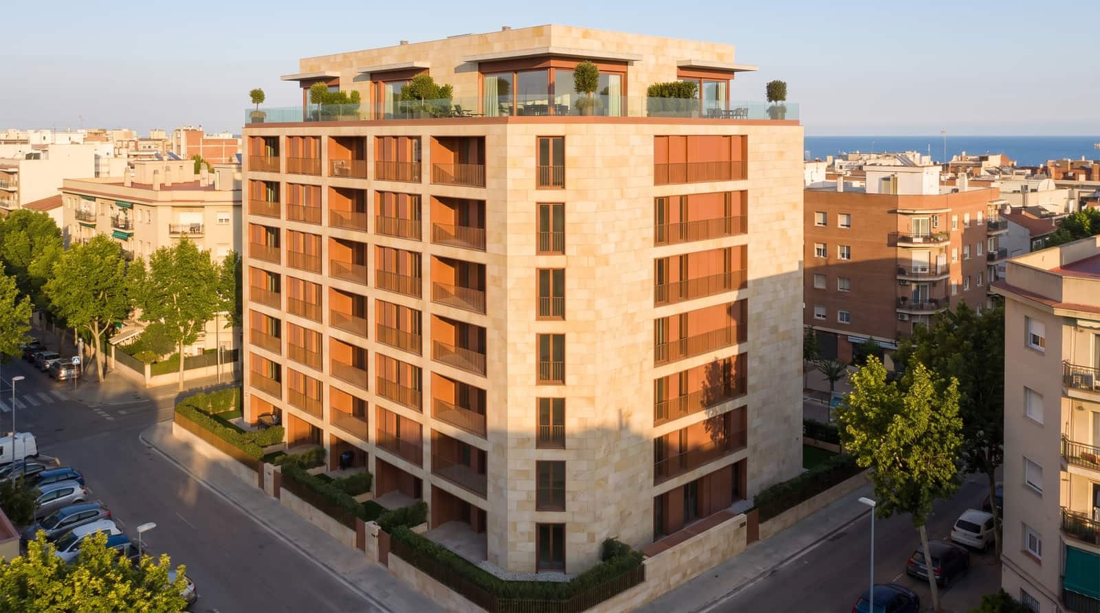 A stone-clad residential building on a Mediterranean street corner, the sea visible on the horizon.